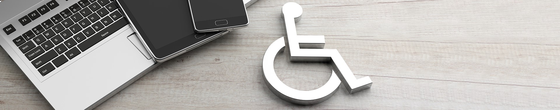 Ein Laptop, ein Tablet, ein Smartphone und das Symbol eines Rollstuhls liegen auf einer Tischplatte.